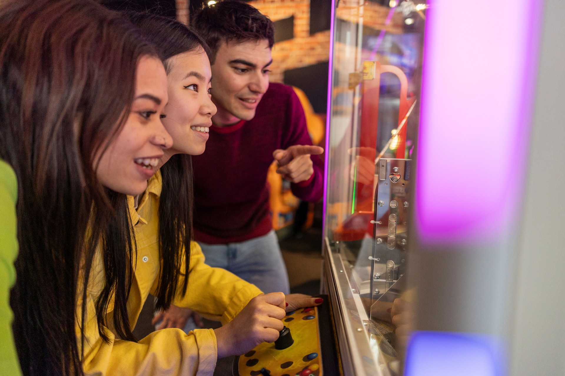 Friends Excitedly Playing Claw Machine at Arcade. Group of young friends eagerly playing a claw machine, focused and excited in a colorful arcade setting.